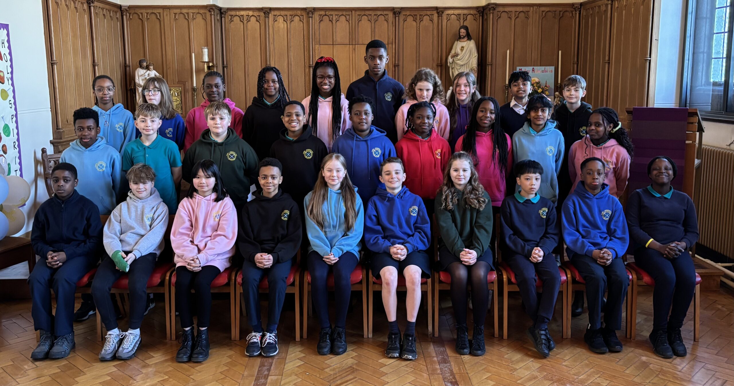 Three rows of pupils sitting or standing in various colours of uniform with wooden panelling in the background.