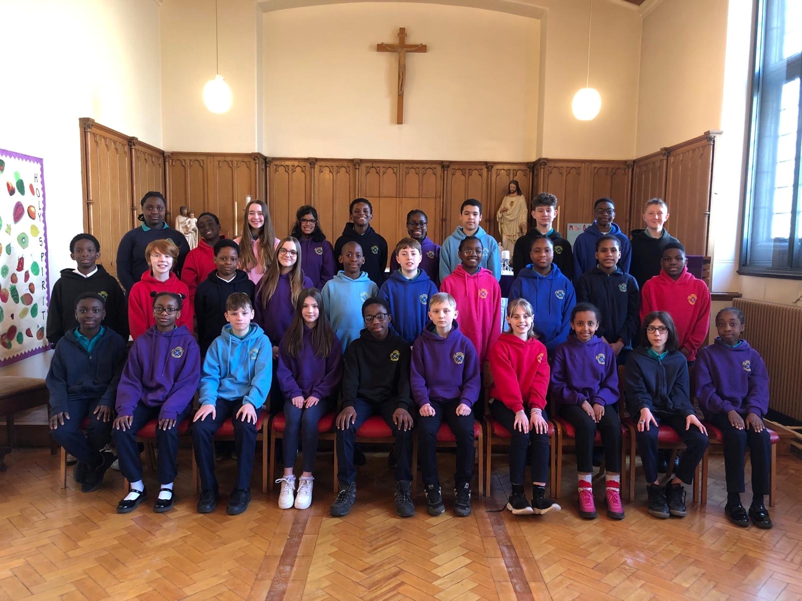 Three rows of pupils sitting or standing in various colours of uniform with wooden panelling and a cross on a white wall in the background.