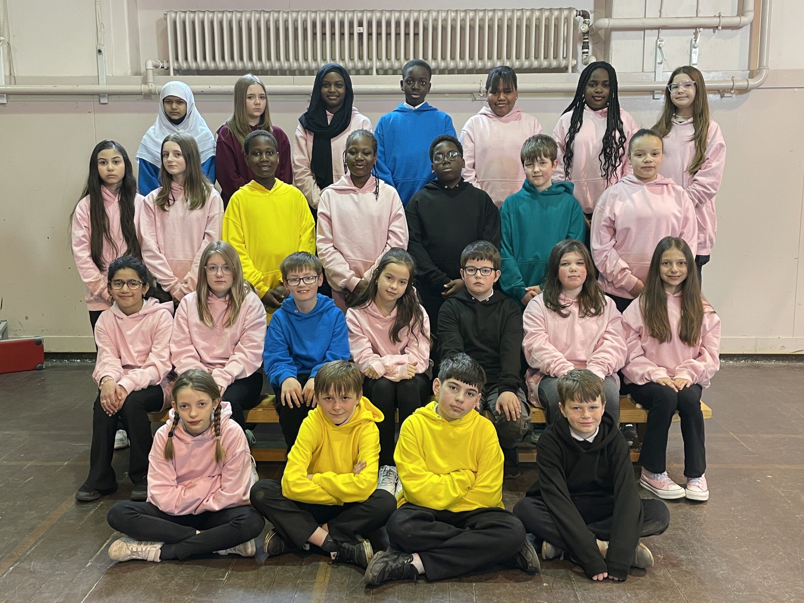 Four rows of pupils in a school hall or gym, mainly in pale pink or yellow tops.