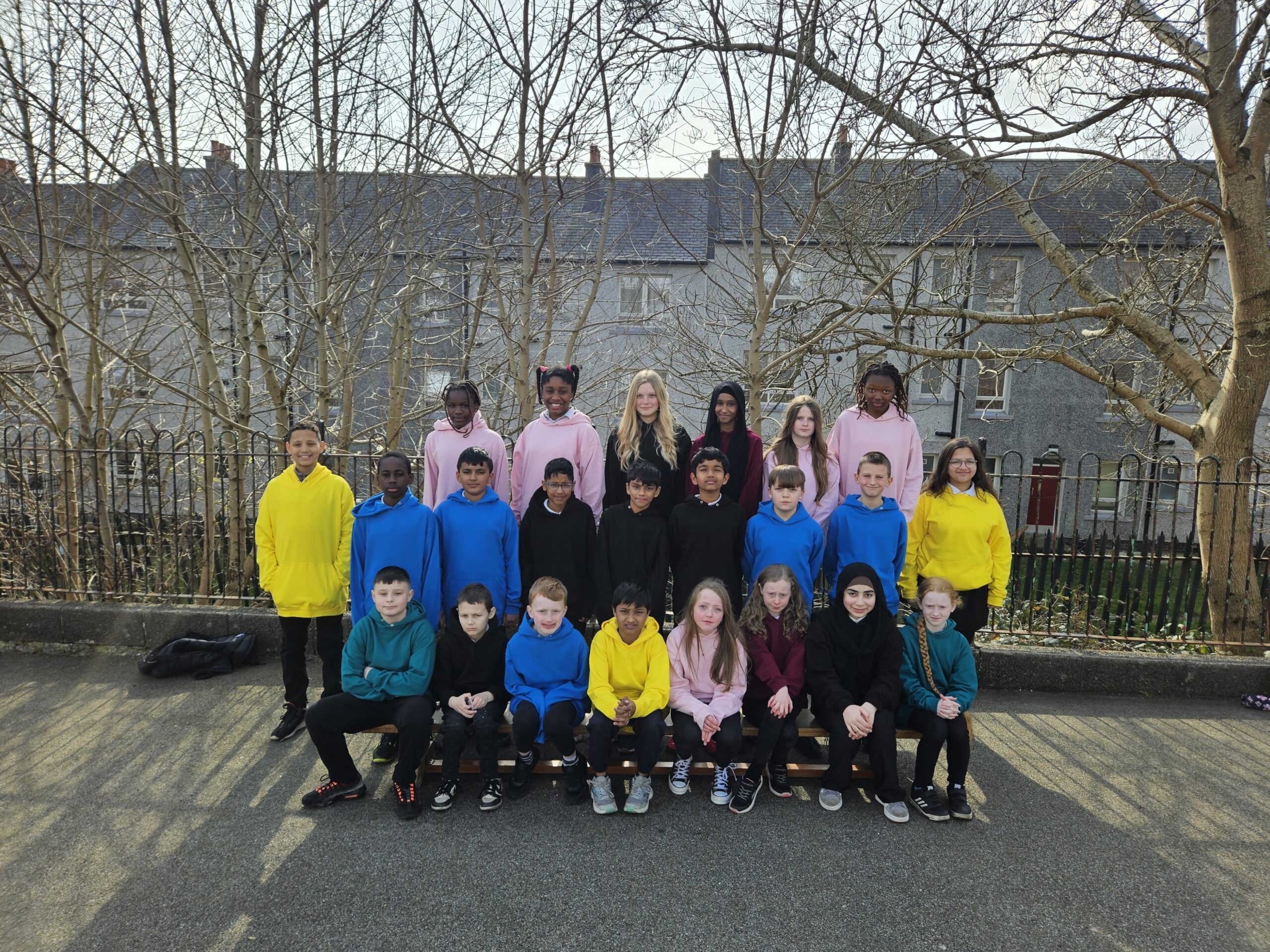Three rows of pupils standing and sitting on benches in a playground with trees and houses behind them, in various colours of school tops.