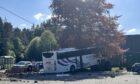 The coach rests against a tree - and on top of a c car that had been parked in the car park. A stone wall, which the bus punched through, is to the left. Debris can be seen scattered across the car park in Dinnet.