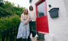 A couple with their black and white collie on the steps of a white cottage with a red door, smiling at the camera.