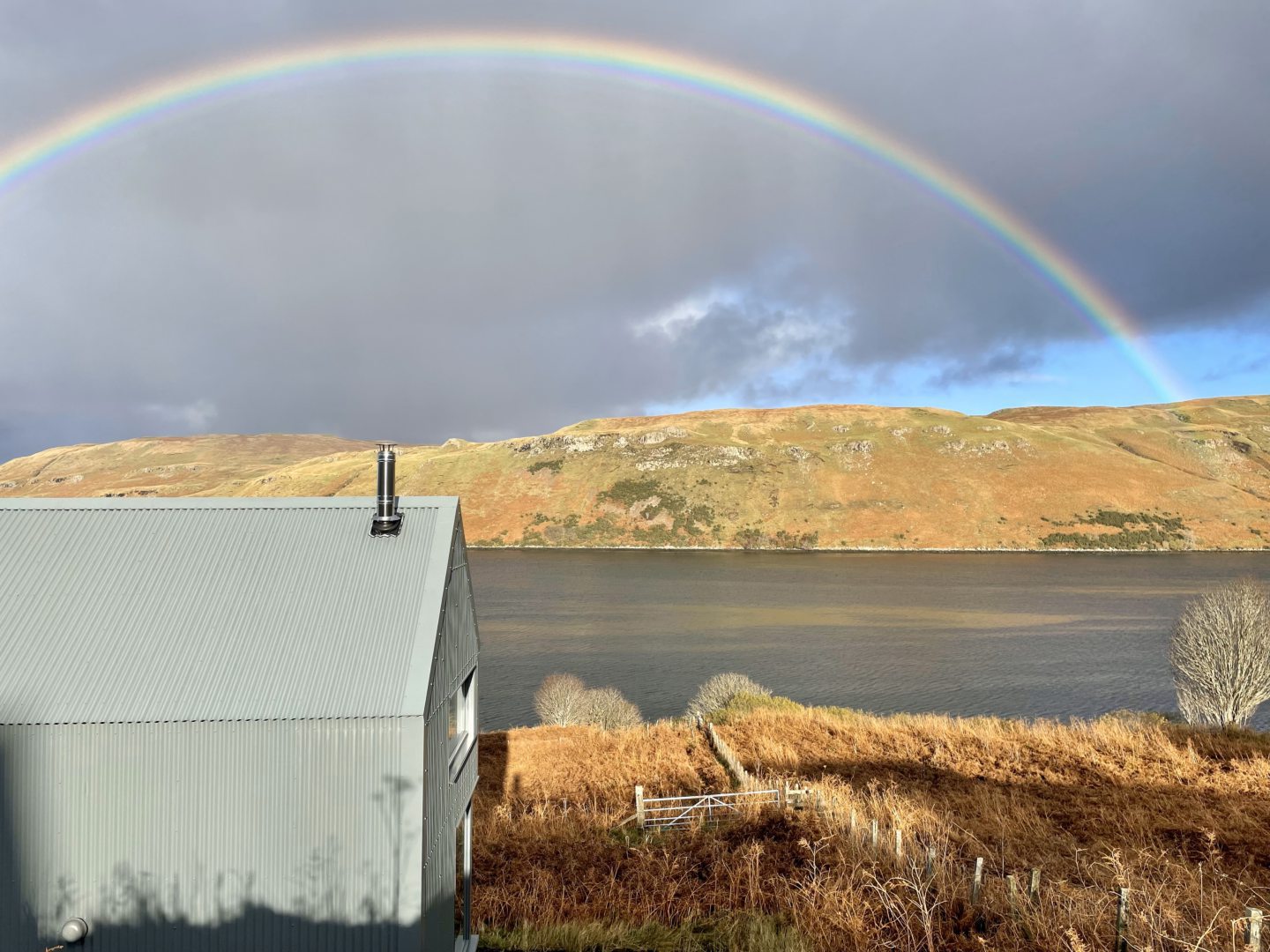 Scotland's Home of the Year's Skye tin-clad cabin in the foreground, with the loch and a rainbow behind.