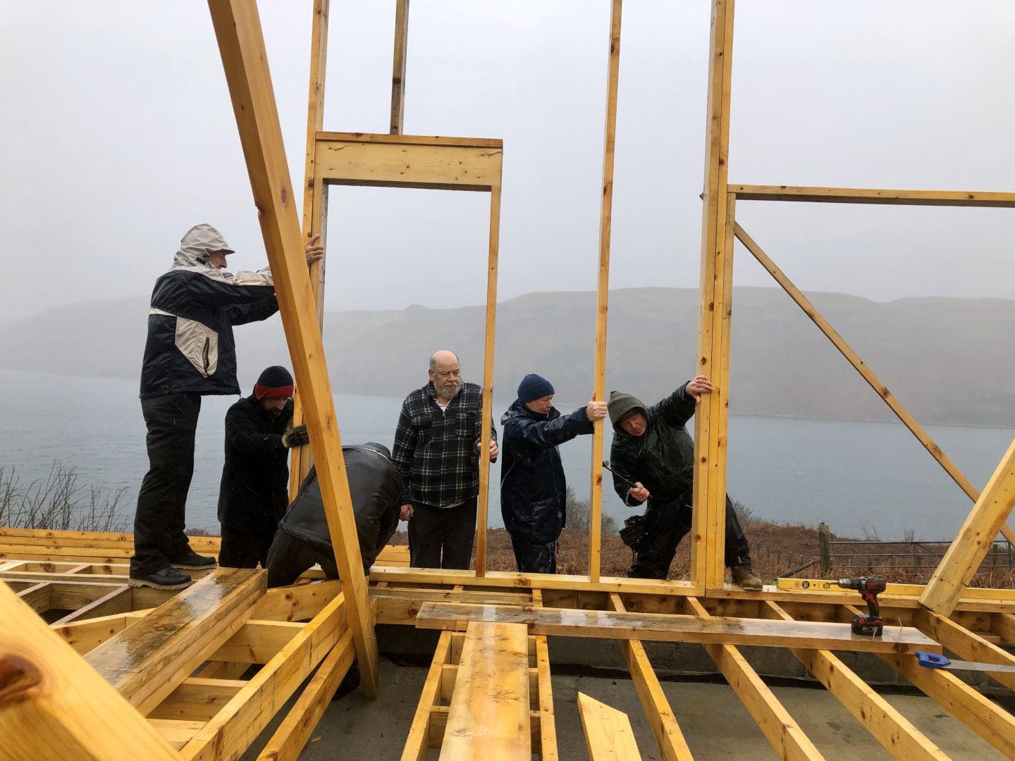 Six people in outdoor clothes helping to erect the wooden frame of a house in the rain.