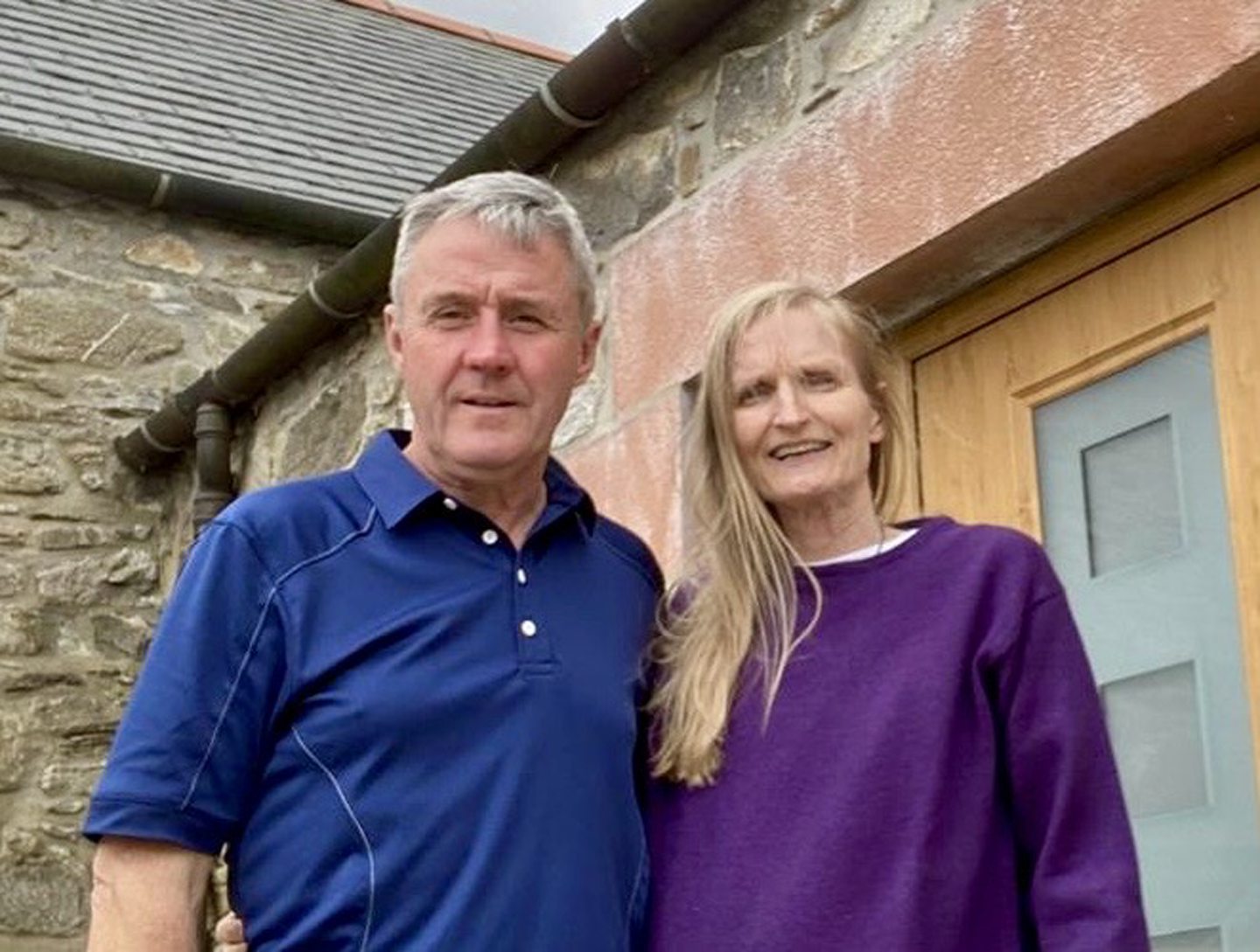 A husband and wife standing next to each other, both in blue tops. She has long blonde hair and he has short grey hair. They are in front of a farm building.