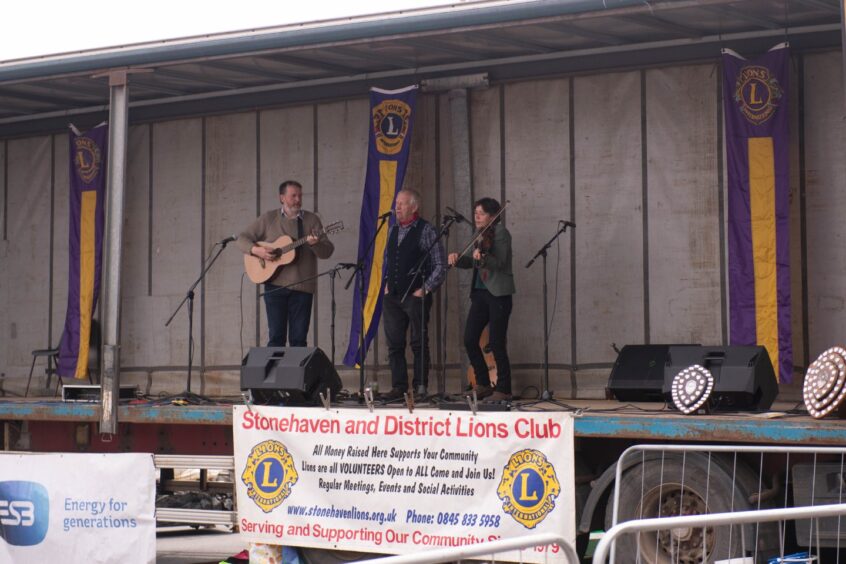 A guitarist, violinist and singer on stage. There is a sign saying "Stonehaven and District Lions Club".
