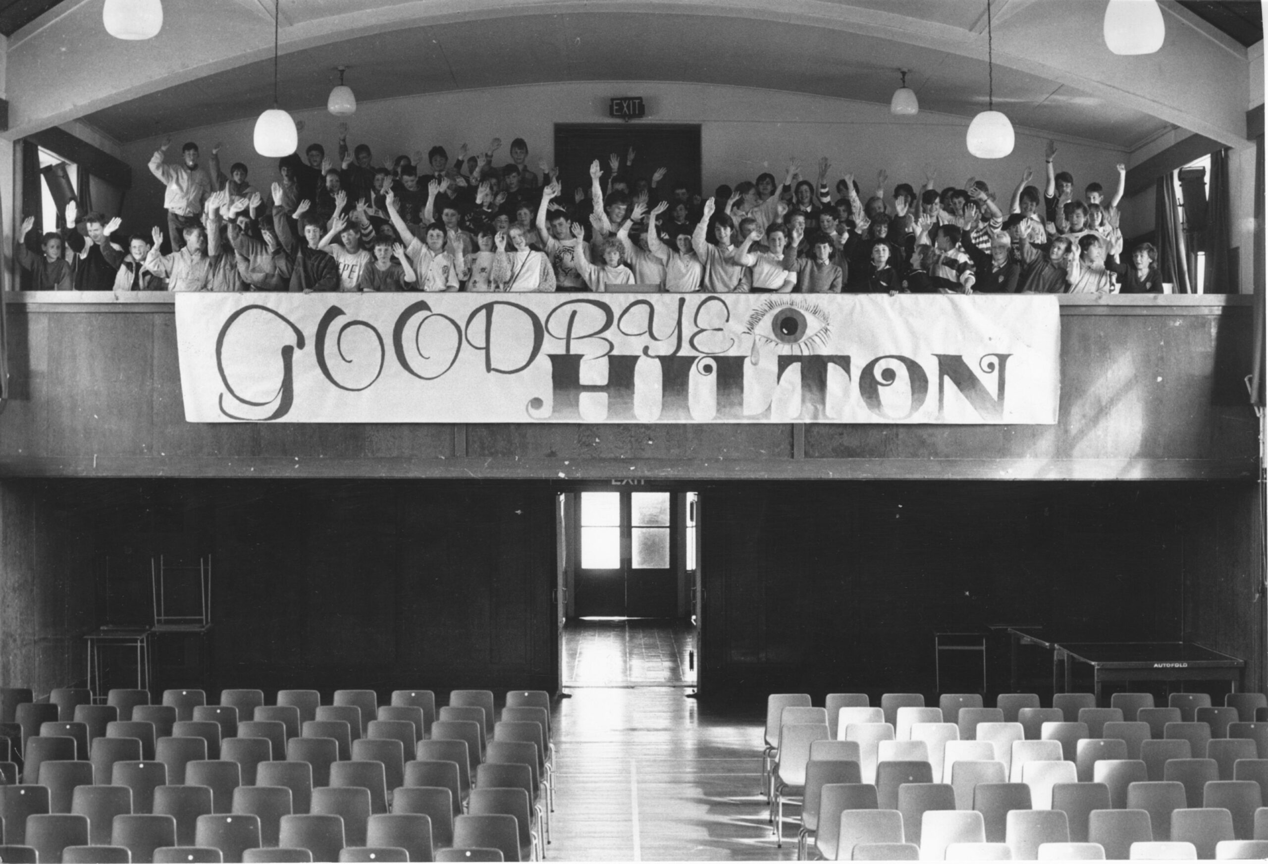 Pupils in the upper balcony waving to the camera with a sign saying "Goodbye Hilton".
