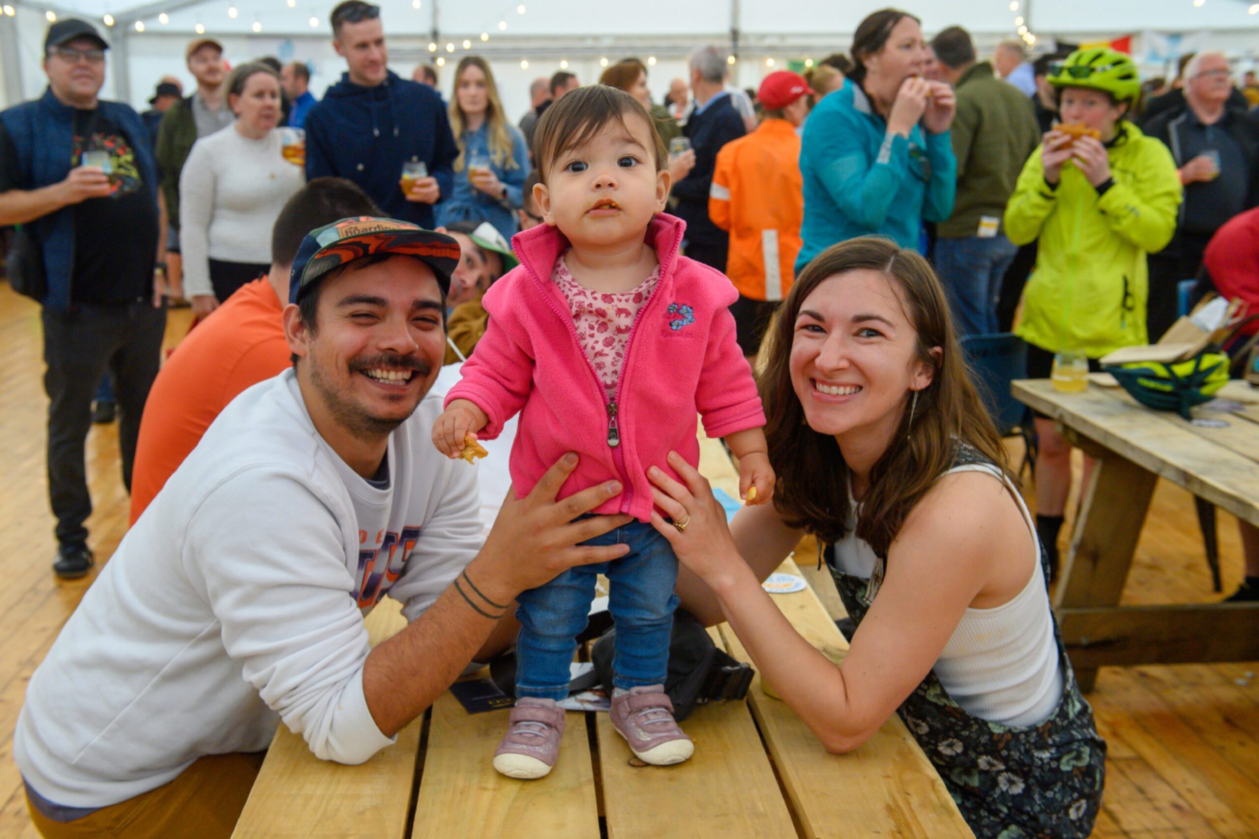 Two adults sit at a picnic table and a toddler stands on it
