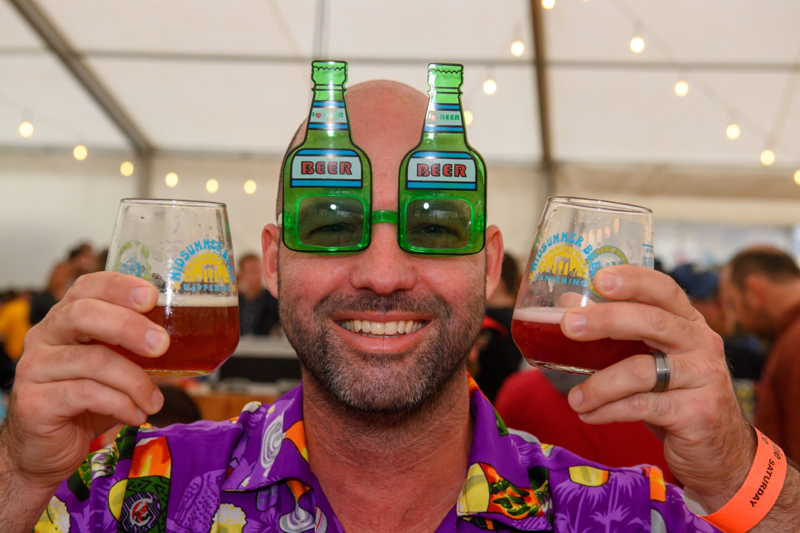 A man wearing glasses that look like beer bottles, holding a glass of beer in each hand