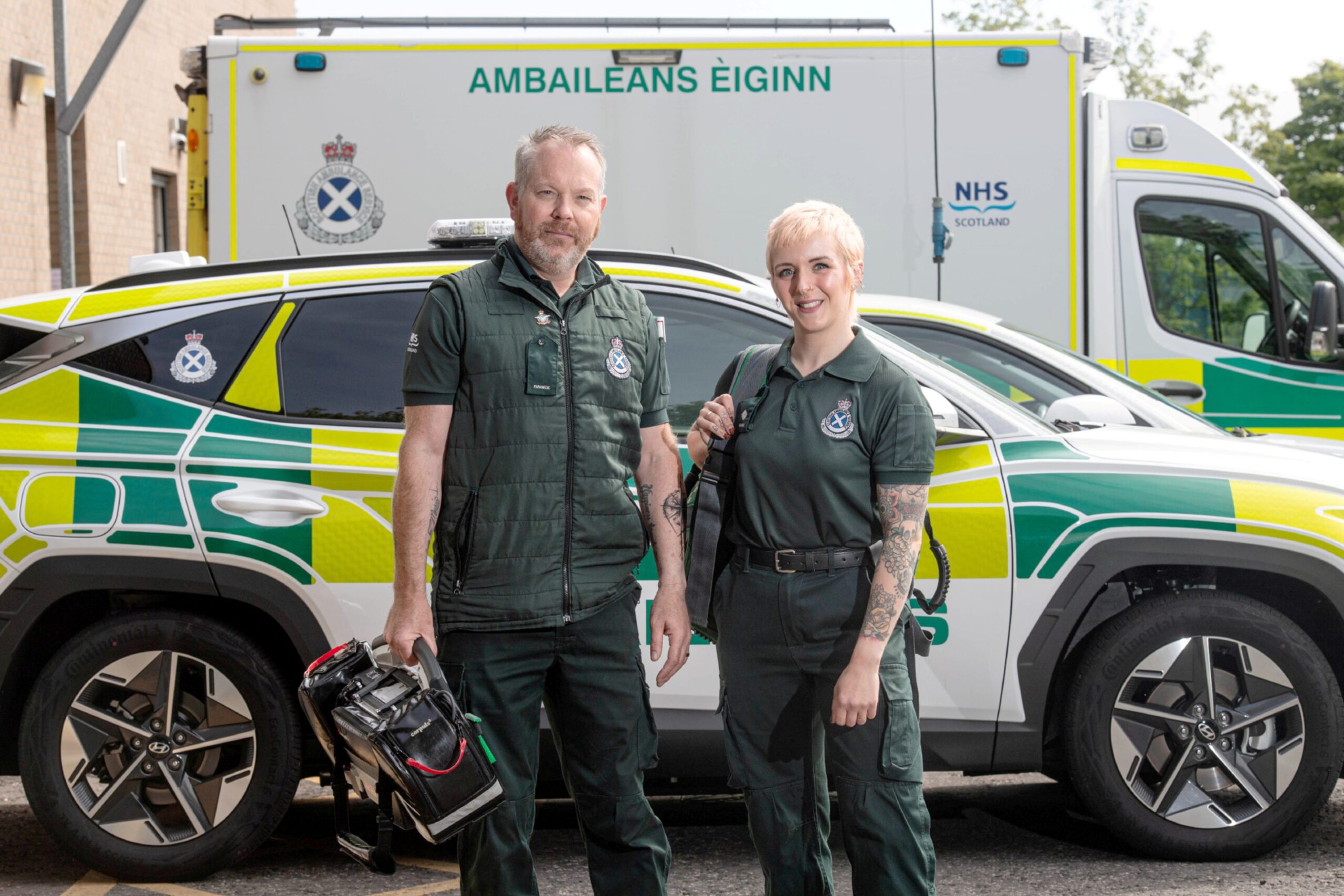 Paramedics Stuart and Sarah Houghton in uniform. There is a rapid response car and an ambulance in the background.