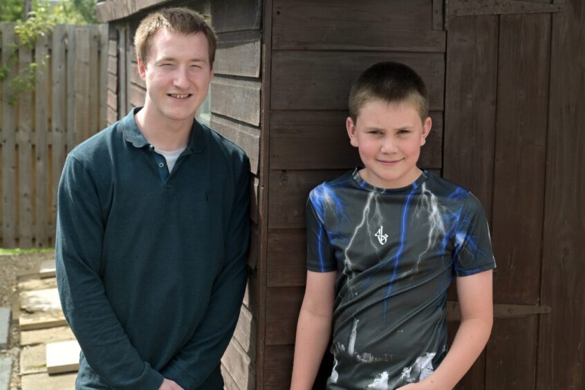 An older boy in a dark top with a collar and a younger boy in a dark T-shirt with a pattern, leaning against a shed and smiling.