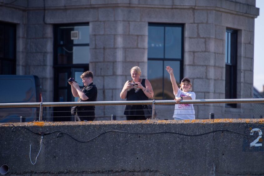 Three people watch on over the harbour wall as boats start to arrive.