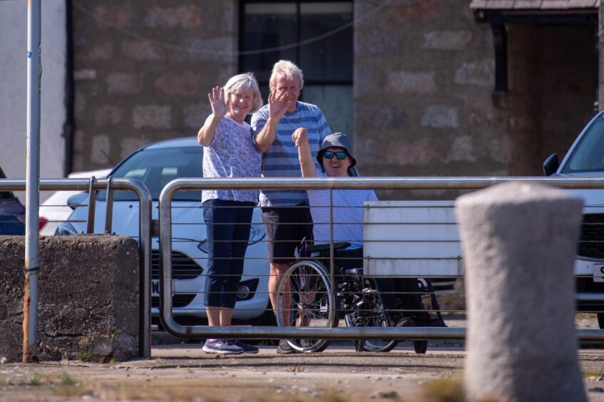 Three people by the harbour wall wave to the arriving ships.