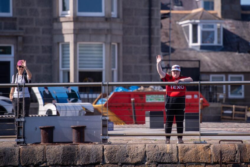 Two people stand by the harbour wall to wave at the ships as they arrive.
