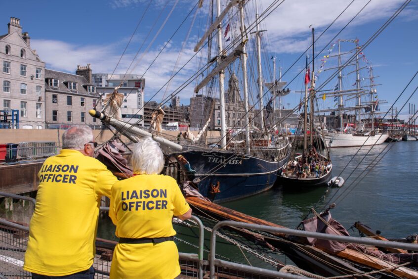 Two liaison officers in yellow shirts overlooking tall ships, including the Thalassa, docked in Aberdeen Harbour before the Tall Ships event.