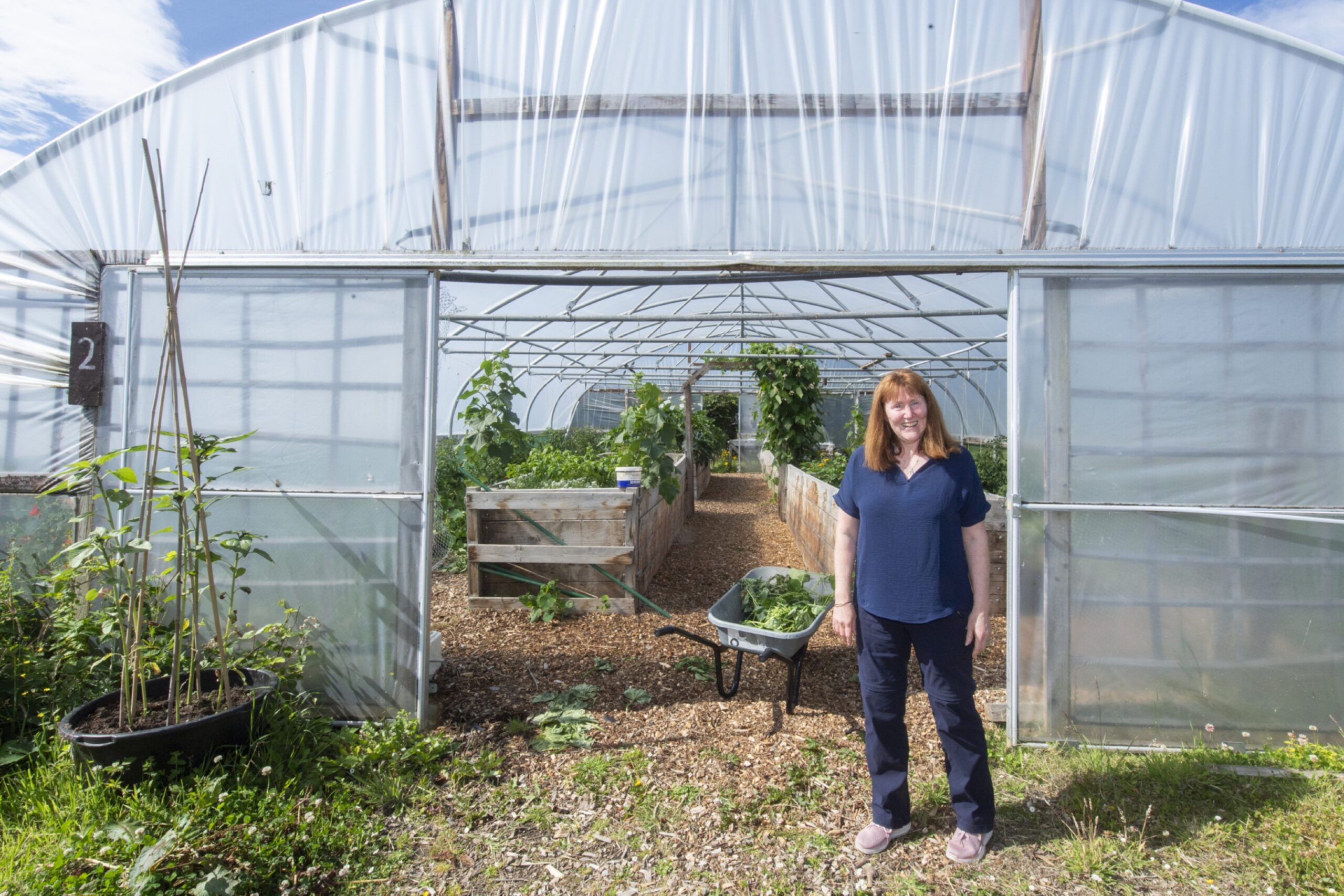 A red-haired woman in a blue tunic-type top and dark trousers, standing in front of a large greenhouse