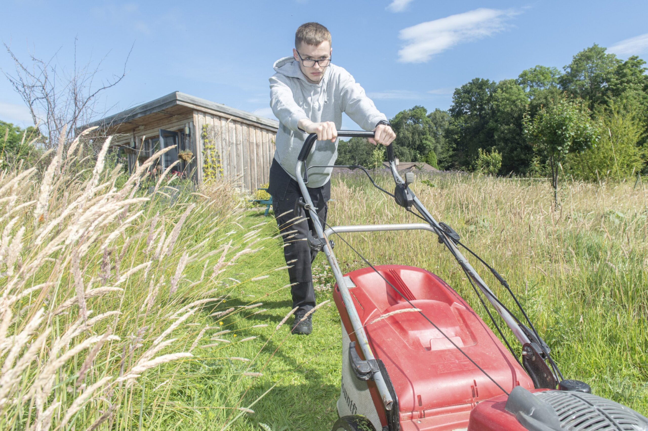 A young man in glasses in a light grey hoodie and dark trousers pushing a red lawnmower