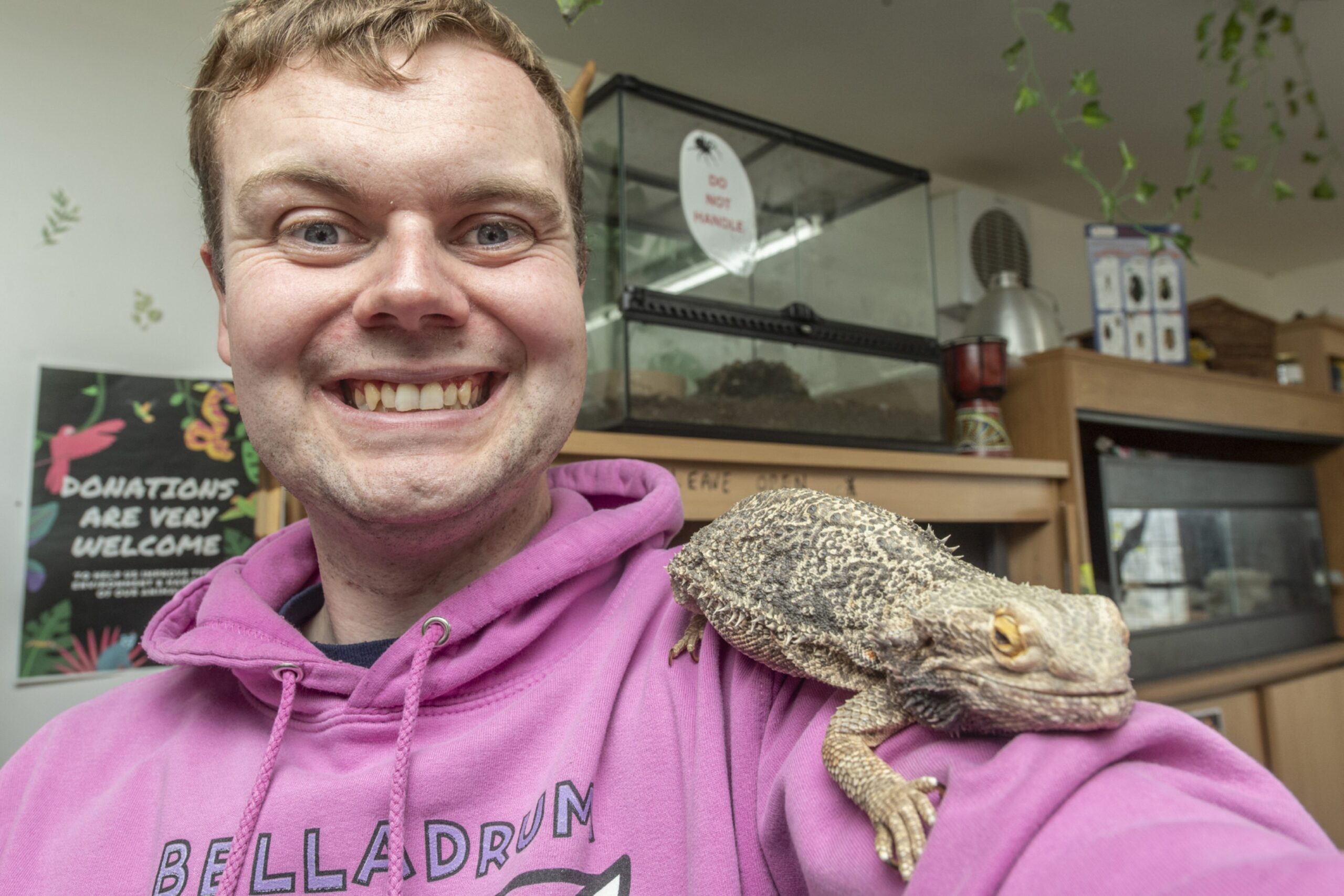 A young man in a pink Belladrum hoodie, smiling at the camera with a reptile on his shoulder