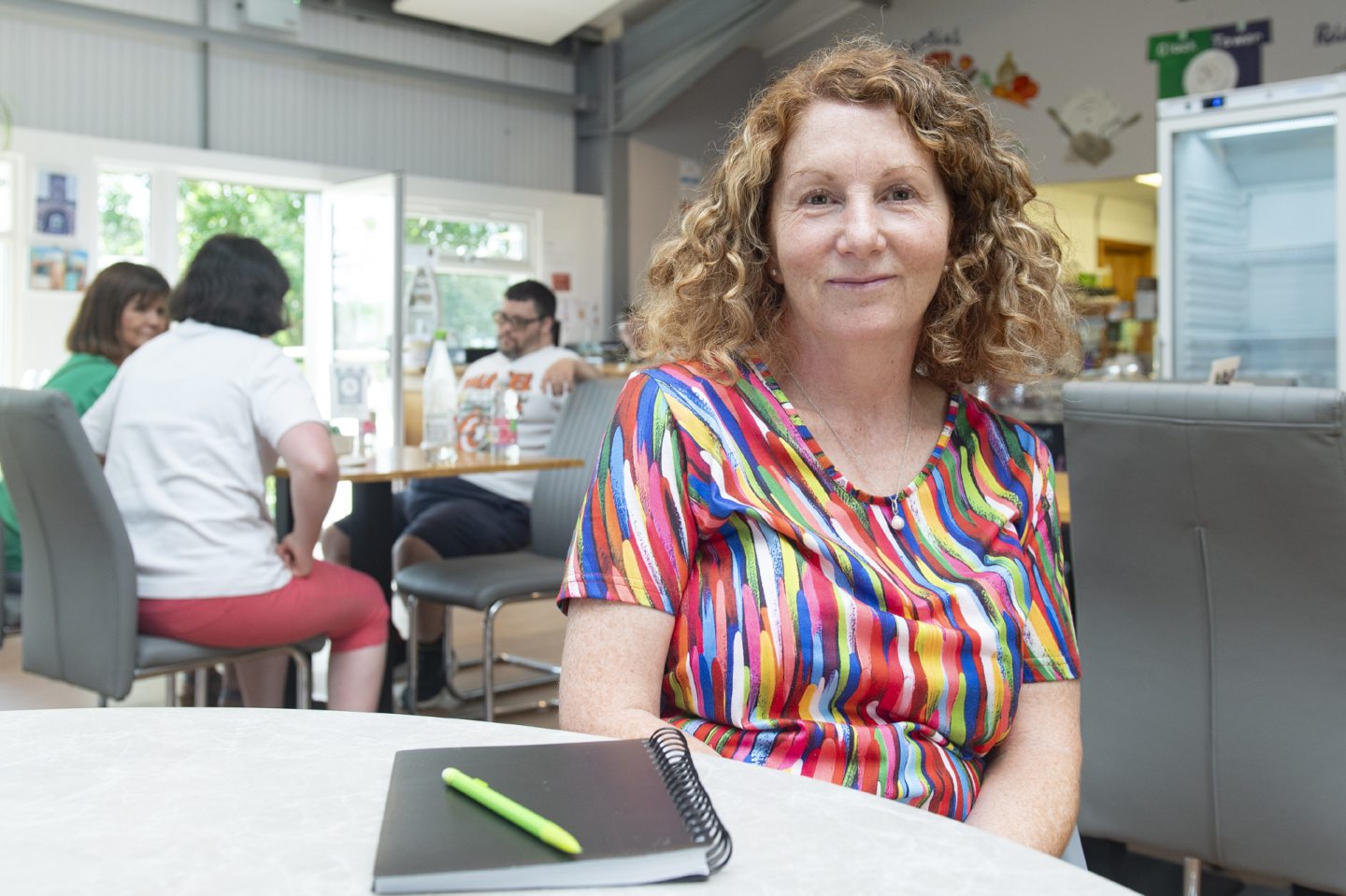 A lady with long reddish blonde hair in a multi-coloured top, sitting at a table and smiling at the camera