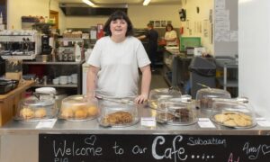 A young woman with dark hair in a white T-shirt behind a busy cafe counter, smiling at the camera.