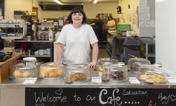 A young woman with dark hair in a white T-shirt behind a busy cafe counter, smiling at the camera.
