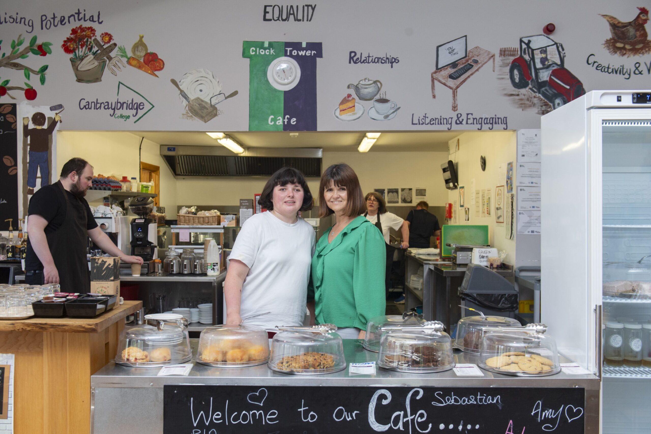 A young woman in a white T-shirt and her mum next to her in a green top, both behind a cafe counter