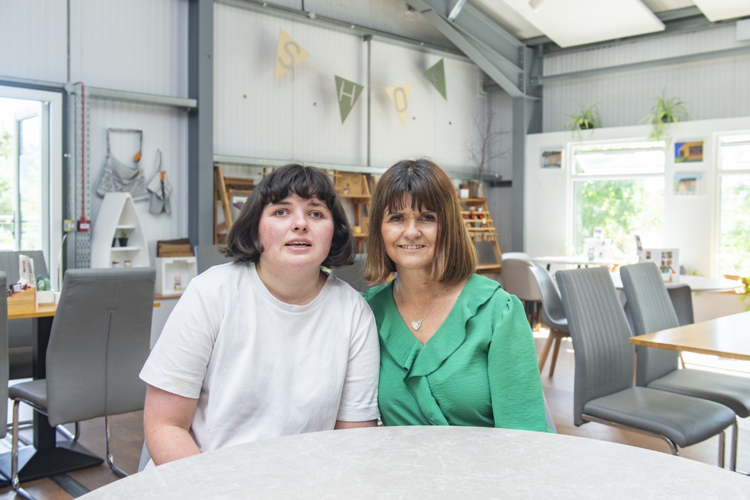 A dark-haired young woman in a white T-shirt and her brown-haired mum in a green blouse, sitting close together at a table.