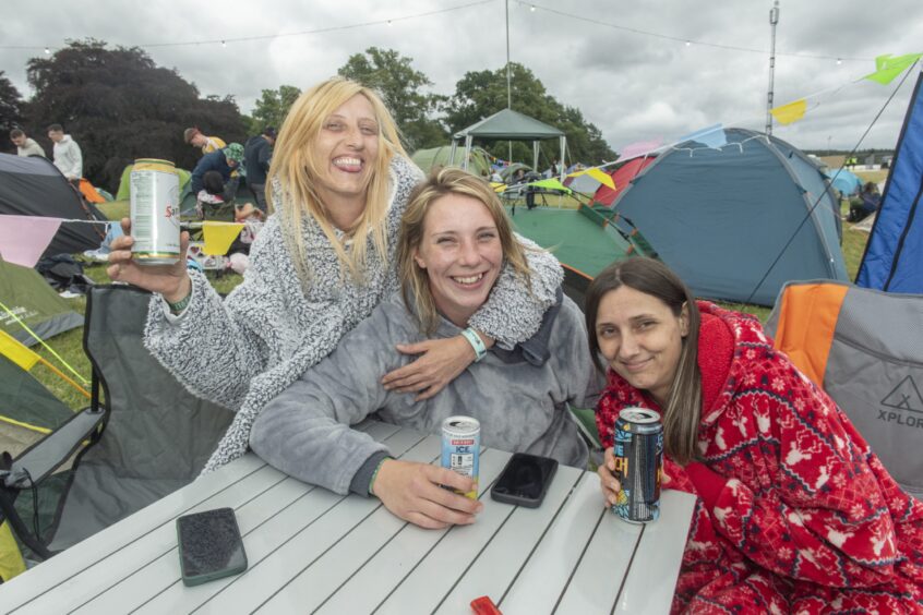 Three young women in furry sleepwear enjoy a tin of beer at a picnic table near the tents.