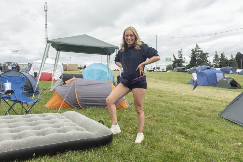 A young woman in shorts and pink shades uses a foot pump to put air into an airbed which is sitting out in the middle of a field with a small one-man tent behind.