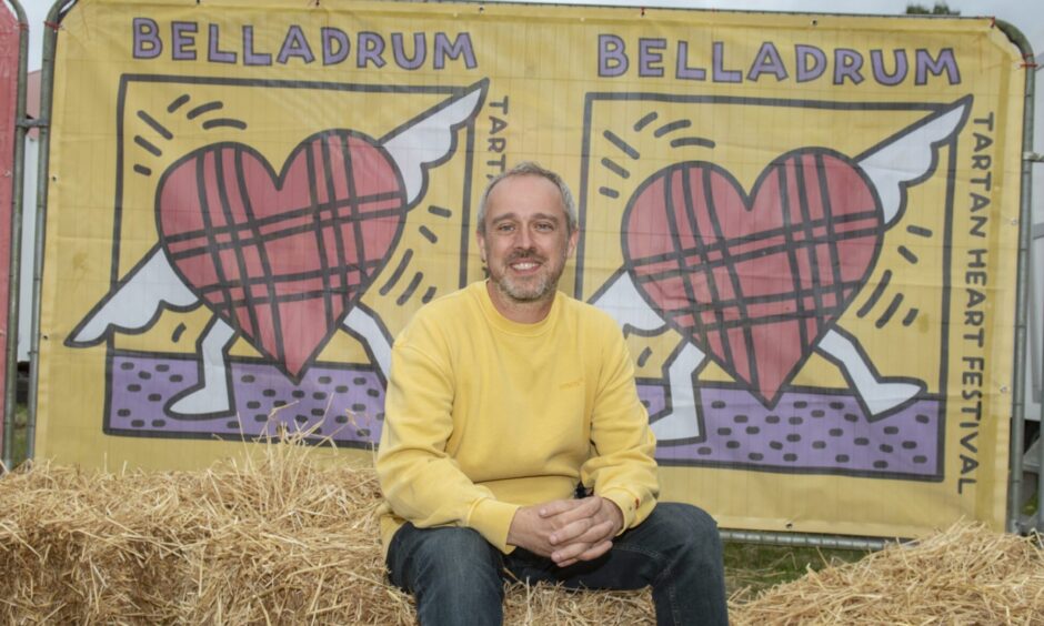 A man in a yellow sweatshirt sits on hay bales in front of the Belladrum Tartan Heart festival banner.