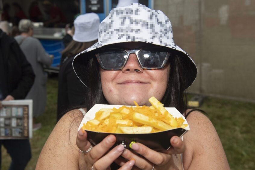 A youngsters in a bucket hat and sunglasses grins as she holds a box of chips and cheese.