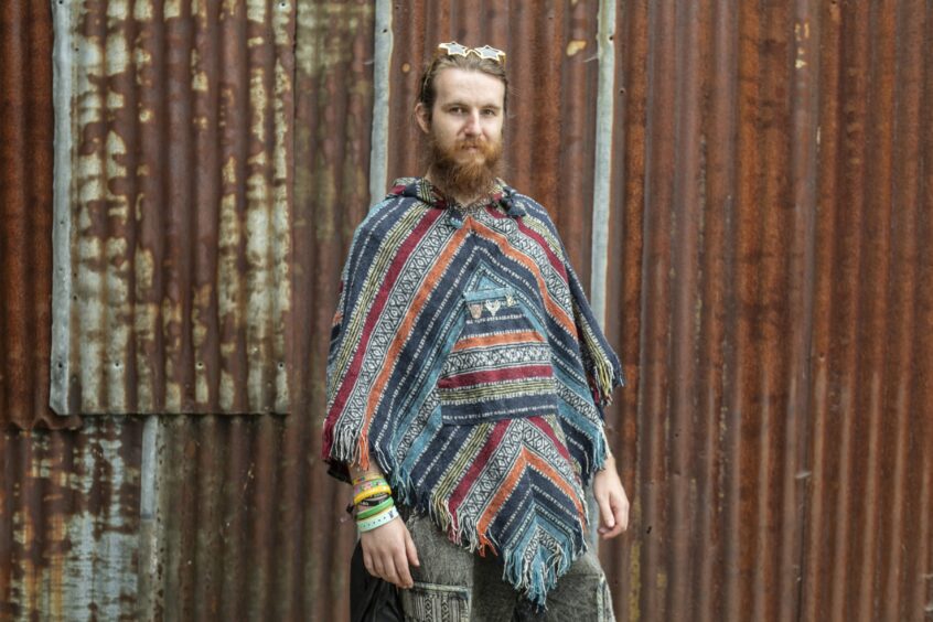 Man in striped poncho, rubber charity wristbands and star-shaped glasses stands in front of a rusted corrugated iron wall.