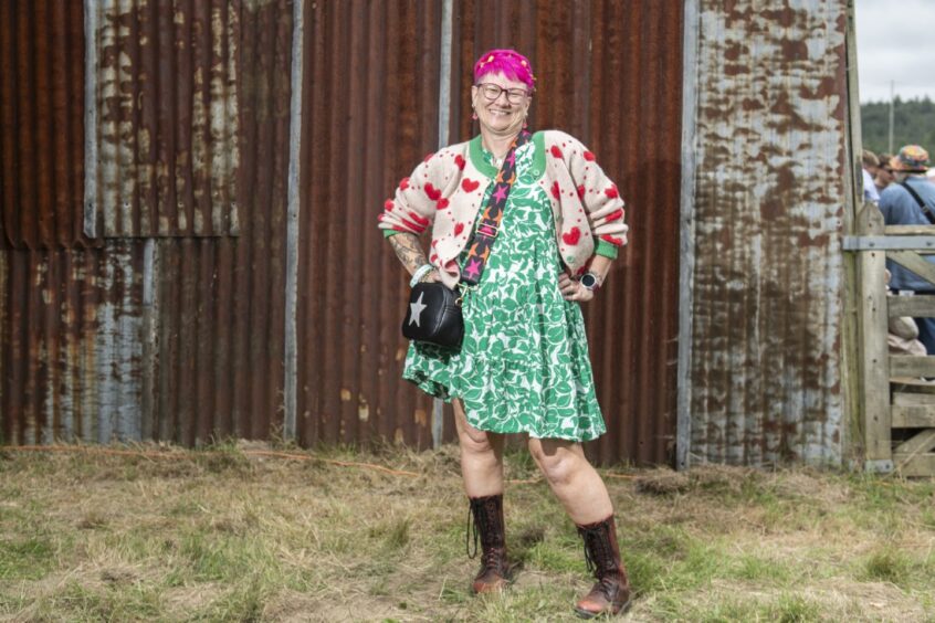 A woman with pink hair wears a green smock dress, pink cardigan with red hearts and handbag covered in stars.