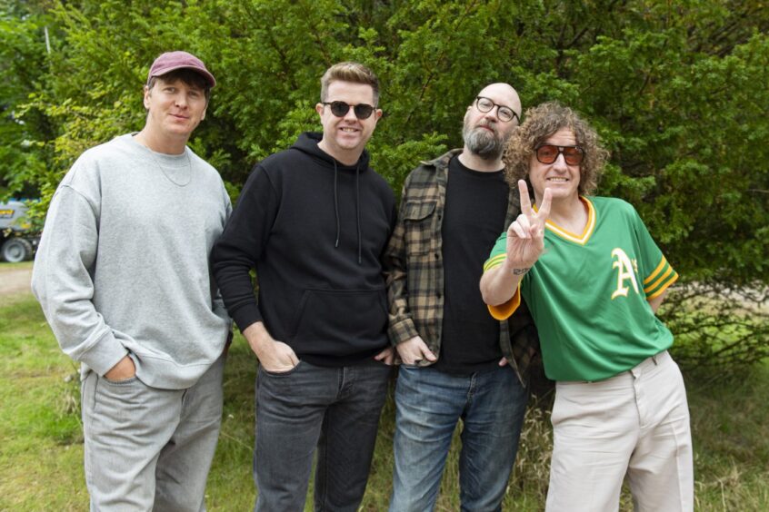 Four men in jeans and sweatshirts stand with their hands in their pockets, the one at the front making the Victory sign.