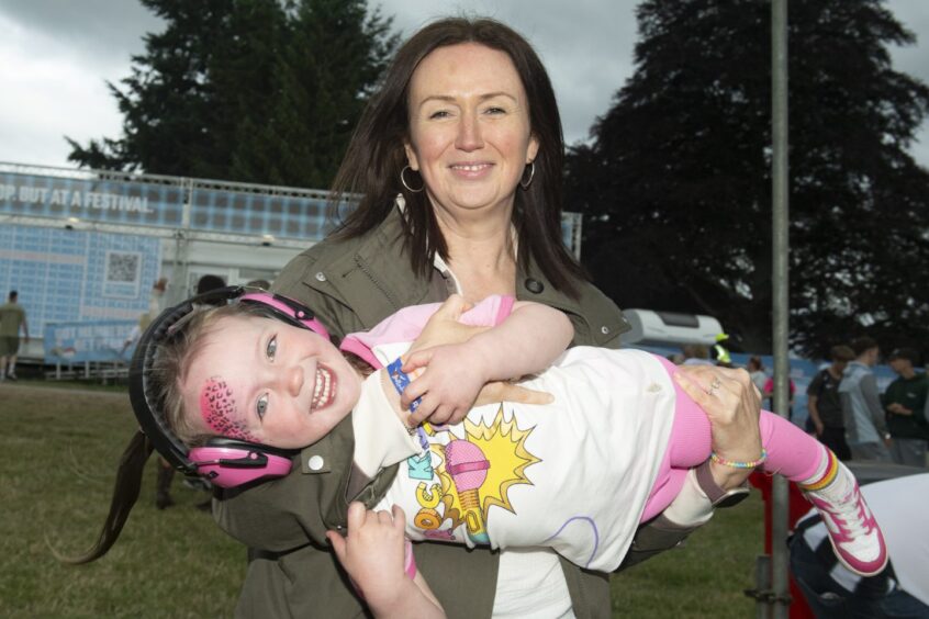 A young festivalgoer in her mum's arms is dressed in pink leggings, t-shirt and ear defenders with a t-shirt adorned with a colourful microphone.