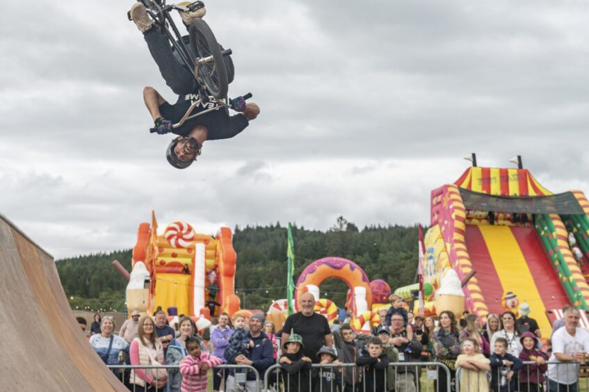A man on a bike turns upside down over a crowd of impressed watchers, with inflatable slides in the background.