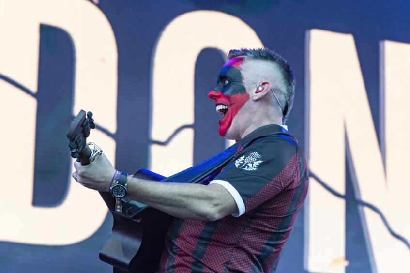 A man with his face painted red and black smiles as he plays the guitar on stage.