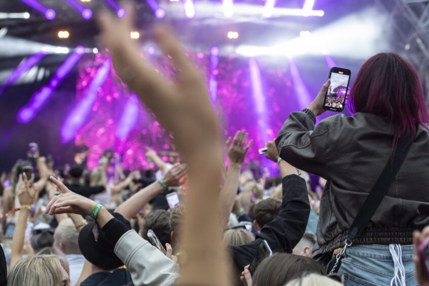 A crowd of hands and a woman watching the stage through her phone screen in front of a pink-lit stage.