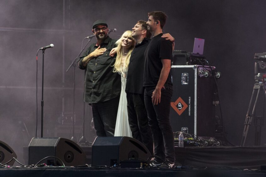 A singer dressed in white and her band in black stand together and acknowledge the crowd.