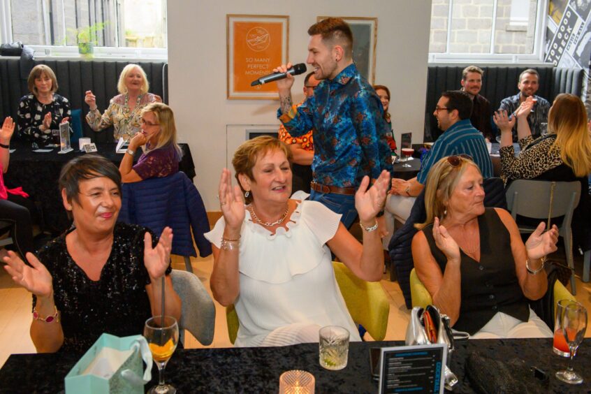 Guests clap along at their tables while a male singer in a patterned shirt performs at That 70s Tribute Show in Aberdeen’s Music Hall.