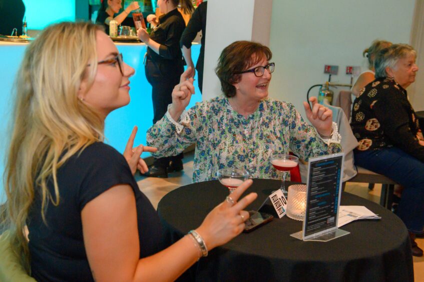 Two women seated at a table making playful dance gestures with cocktails in front of them during That 70s Tribute Show at Aberdeen’s Music Hall.