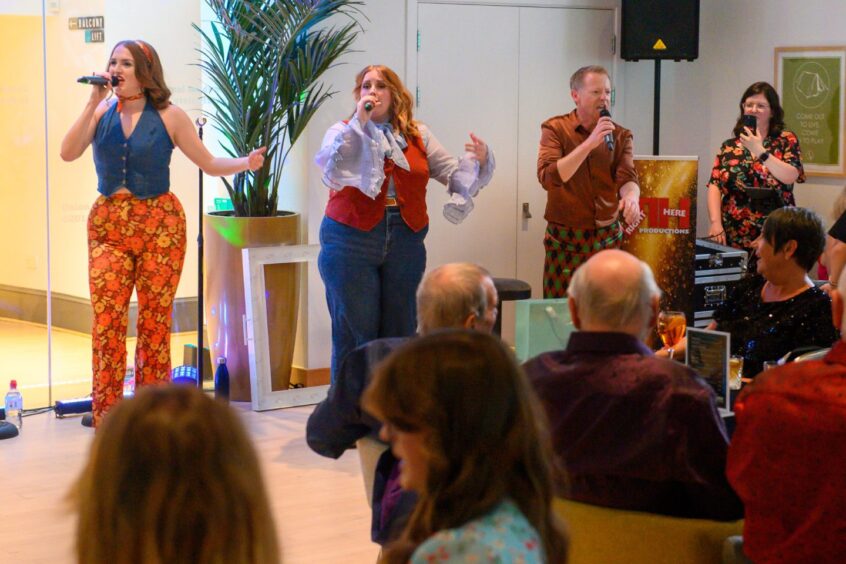 Three singers in colourful 70s outfits perform on stage at That 70s Tribute Show in Aberdeen’s Music Hall, with guests watching from seated tables.