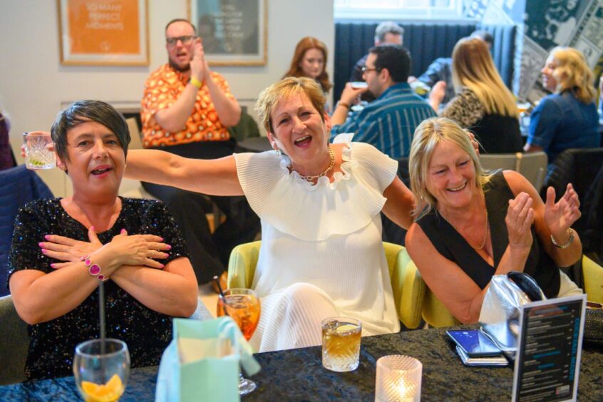 Three women laughing and singing at a candlelit table with drinks during That 70s Tribute Show at Aberdeen’s Music Hall, surrounded by other guests.