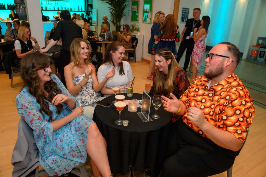 Group of guests laughing and chatting around a candlelit table with cocktails at That 70s Tribute Show in Aberdeen’s Music Hall, one wearing a bold retro shirt.
