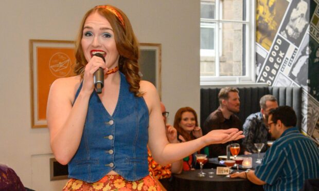 Singer in floral trousers and denim waistcoat entertains guests at That 70s Tribute Show in Aberdeen’s Music Hall, with seated audience enjoying cocktails.