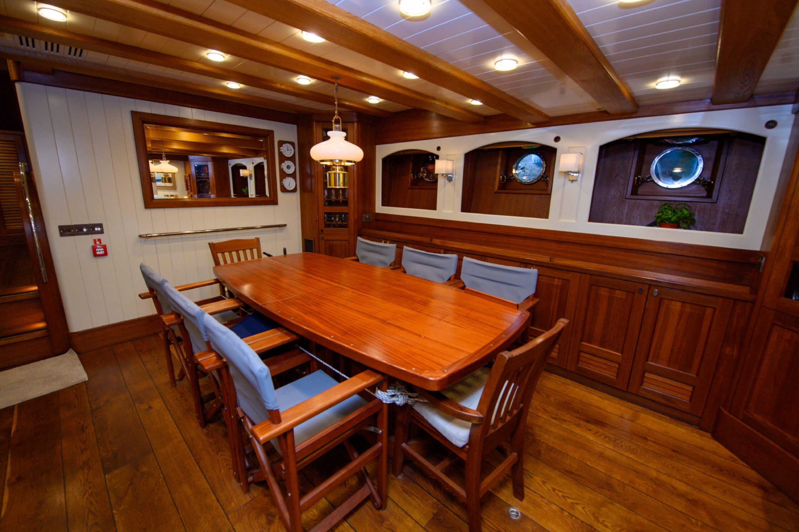 The dining area of the Malcolm Miller, which includes a wooden table with eight chairs lined with blue cushions, three port-holes and a mirror on the back wall.
