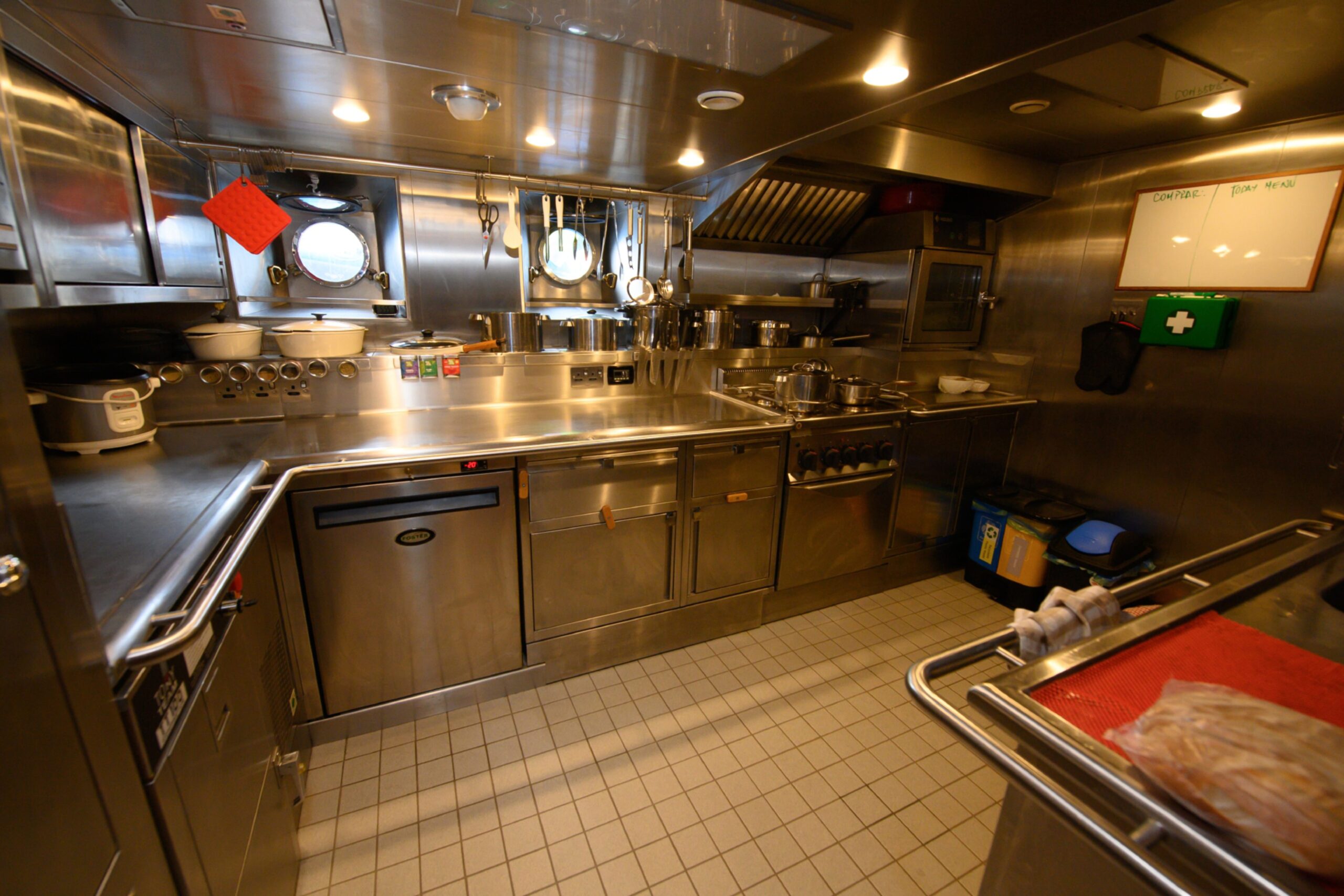 The ship's galley, with shiny metallic cooking surfaces, neatly-organised utensils and a whiteboard on the far wall.