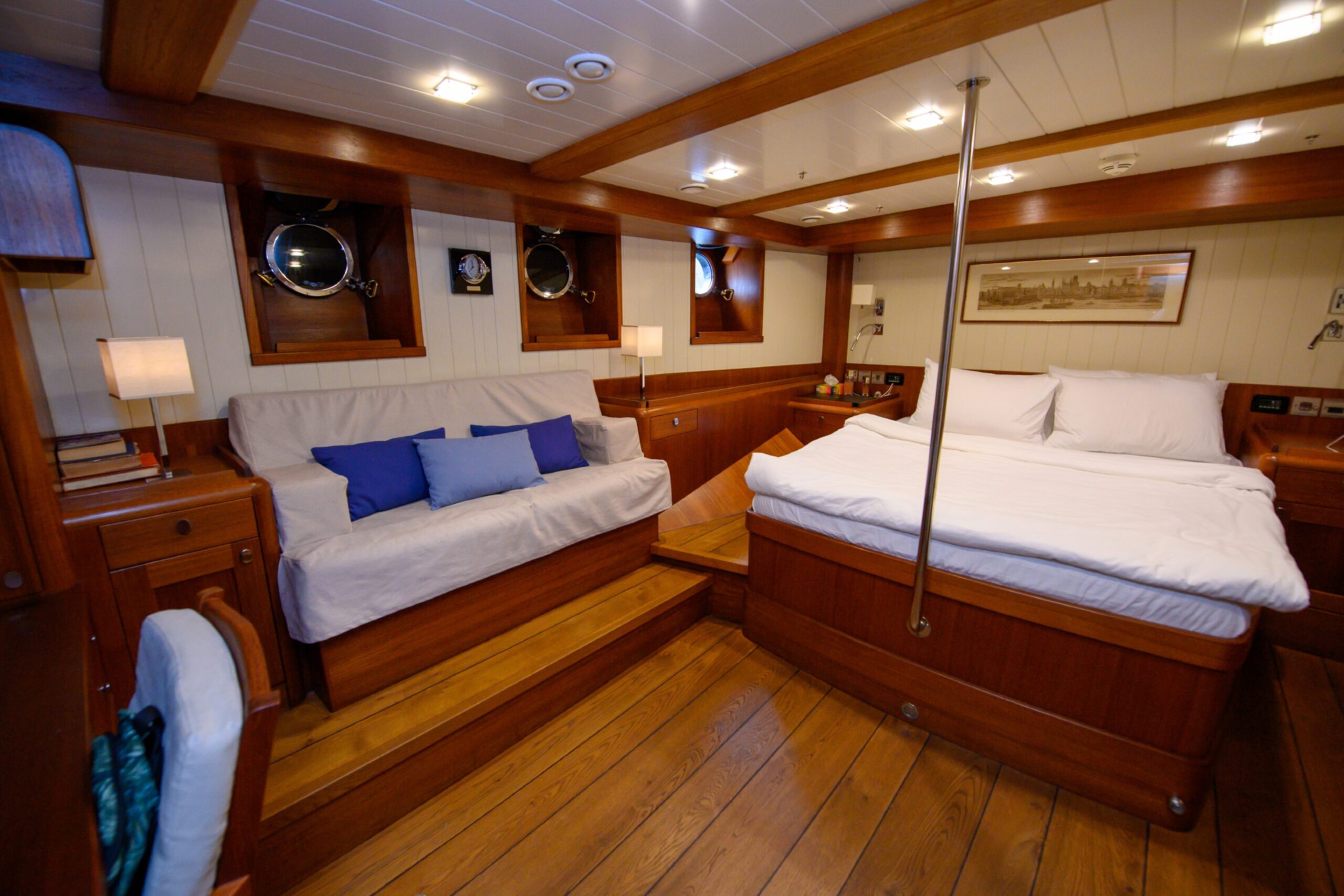 The ship's master bedroom, which includes a large bed with white bedding, a sofa bed with blue cushions, three port-holes, wooden decor and pictures adorning the walls.