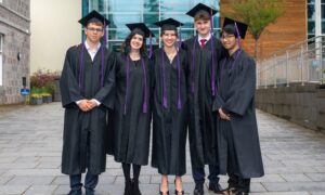 Five students, boys and girls, in gowns and mortar boards, lined up and smiling for the camera