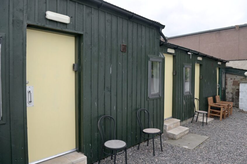 The wooden-clad building. It is painted green with yellow doors. The building has four doors which lead to hotel rooms. There are tables and chairs outside and a gravel driveway. 